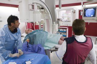 Пациент, лежащ на рентгенова маса до лекар, гледащ изображение на кръвоносните съдове на пациента на екран